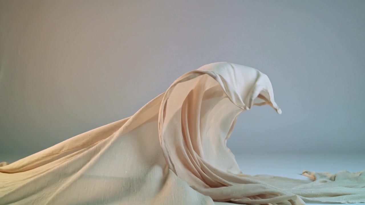 Lifting large beige fabric with hem forming crest and curling on white table from offframe airflow