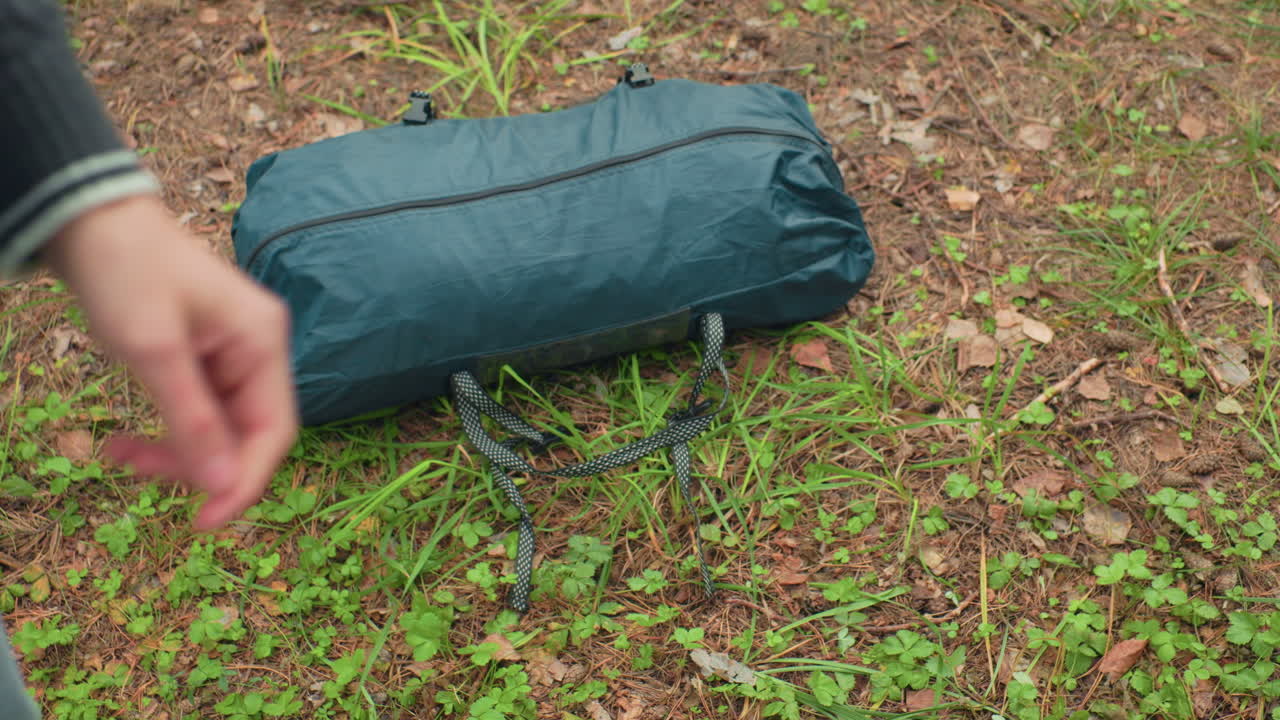 close up partial view of green duffel bag being dropped onto forest ground covered with green vegetation and dry leaves, showing start of outdoor activity or camping preparation in woodland area