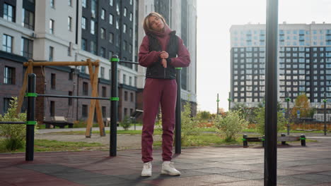 sports woman stands beside outdoor bar under bright sun, rolling hands and neck in slow warmup sequence before workout in urban park surrounded by tall residential buildings and clear sky