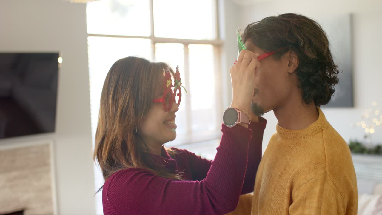 Vielfältiges Paar umarmt sich, rückt die Brille zurecht, trägt eine Smartwatch, drückt die Wange aneinander und lacht zu Hause