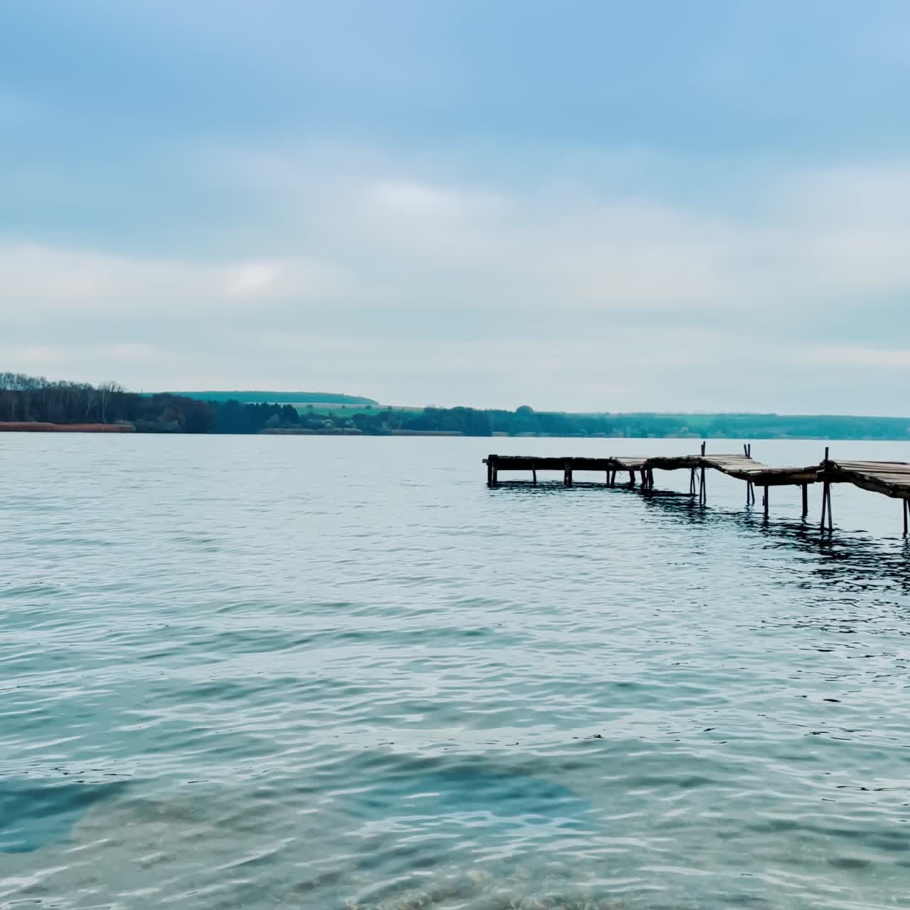 Wooden clumsy bridge going to the river. Bare dark trees at the waterfront on the other side of the river. Blue grey cloudy sky at backdrop