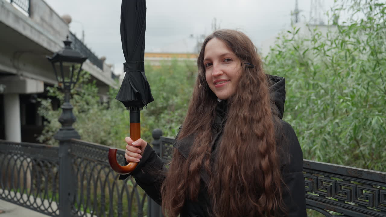 Giovane donna che cammina su un ponte in città. Donna sorridente con i capelli ricci che attraversa il ponte sul lungofiume urbano. Allegra giovane donna con cappotto nero che porta un ombrello lungo il sentiero sul fiume in città.