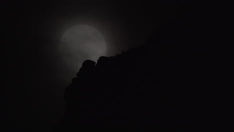 a full moon rises beyond a cloudy night sky and mountain top