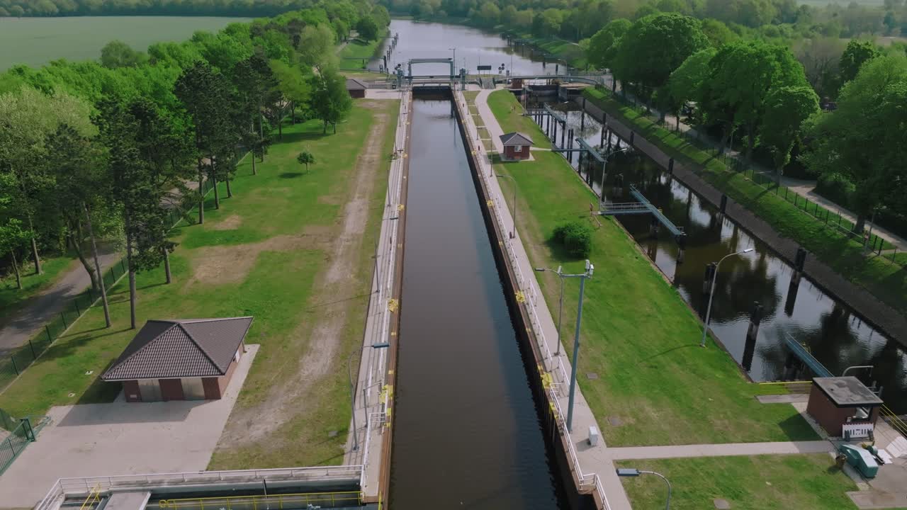Zoom out showing an empty water lock and surrounding canal system with lush greenery under clear skies.