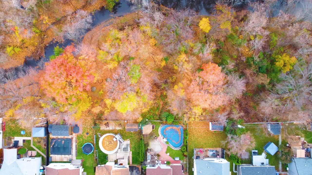 A straight-down drone shot reveals a vibrant fall scene of backyards with pools and sheds, set against a backdrop of a river.