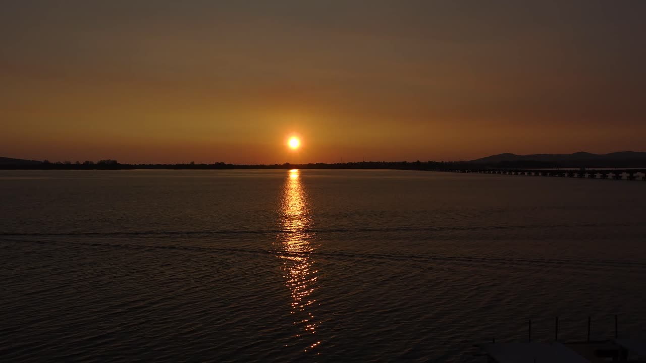 Auringonlaskun aikaviivekuvaus tyynen veden yllä Thaimaan ja Kambodžan rajalla Koh Kongin maakunnassa, hehkuvan heijastuksen, kaukaisen sillan ja lämpimän iltataivaan kera.