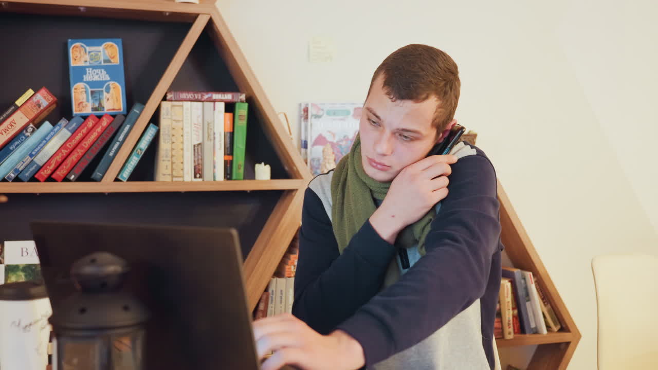 Uomo bianco al telefono davanti al laptop in uno studio luminoso, con una sciarpa verde e un'espressione composta, che gesticola mentre parla. Sfondo con libreria organizzata, tazza di caffè e lanterna sulla scrivania, atmosfera da teleconferenza.