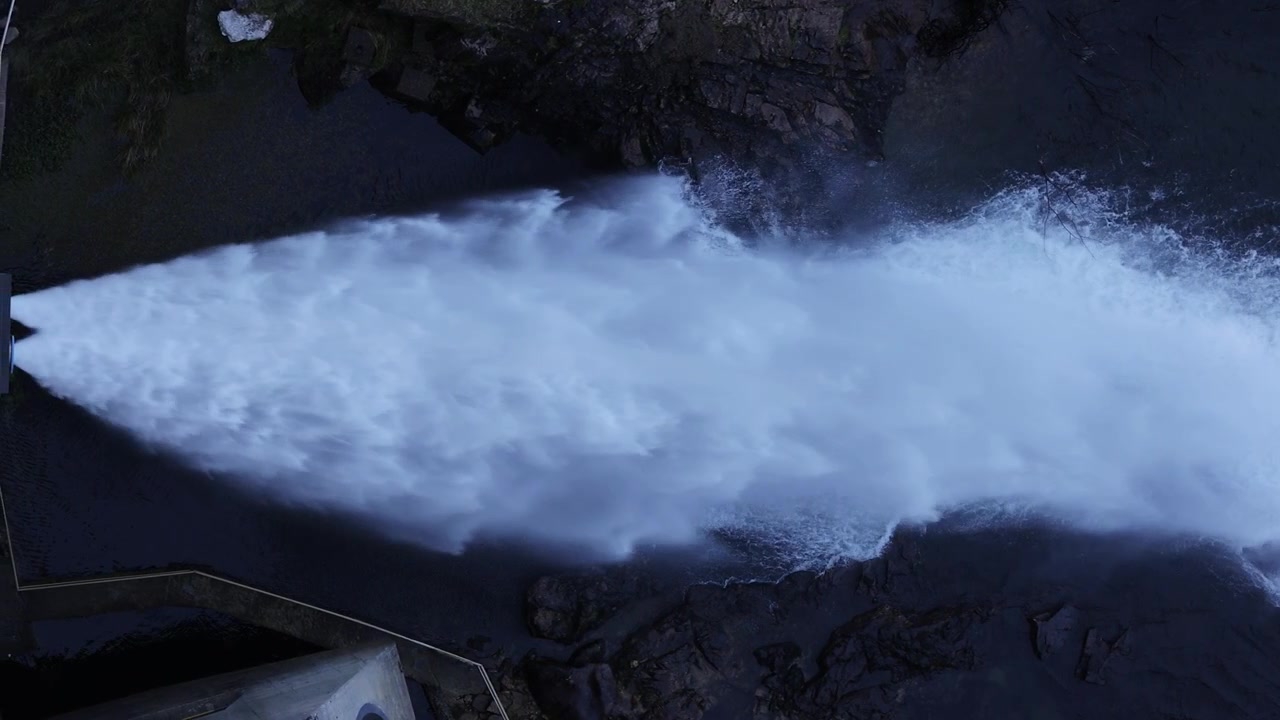Impresionantes imágenes aéreas capturan la potente descarga de agua de una presa hidroeléctrica, golondrinas planeando a cámara lenta sobre corrientes espumosas, hormigón industrial, iluminación natural de la hora azul