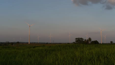 Wind-Turbines-shooting-out-of-a-Farmland-while-the-Sun-is-setting-as-Trucks-Pass-by-making-Clouds-of-Dust,-clean-alternative-energy-in-Thailand-and-mainland-Southeast-Asia