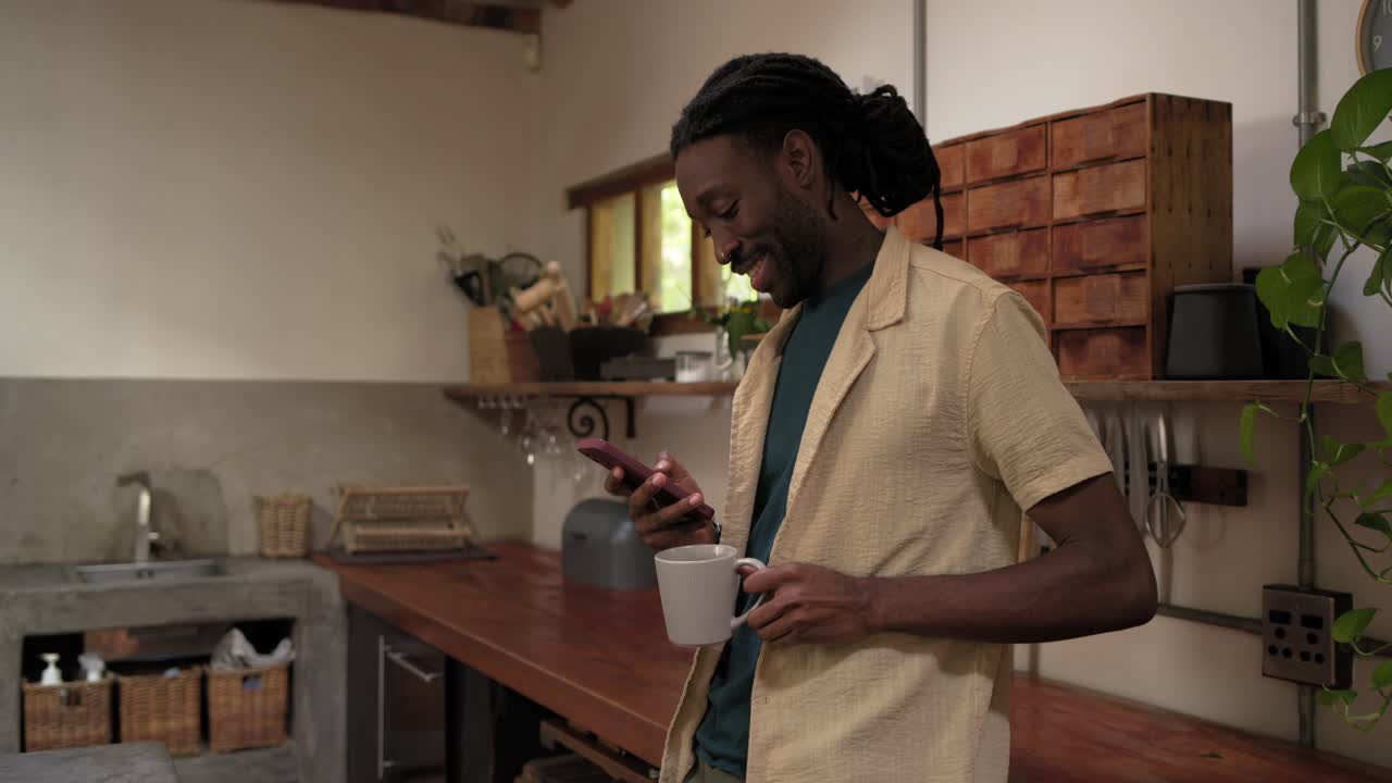 Homme afro-américain en chemise beige, appuyé à un comptoir en bois, consultant son téléphone, sirotant une tasse et lisant des messages.