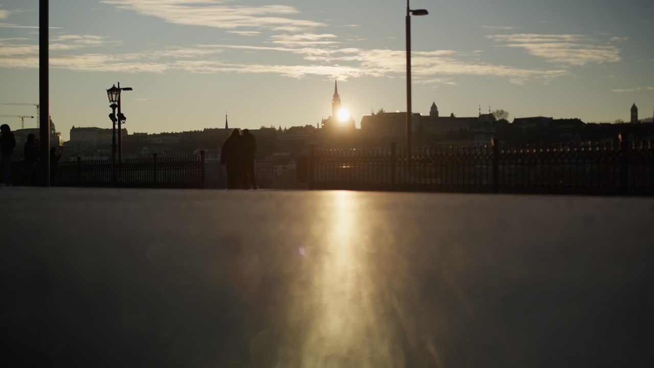Piernas y pies de personas caminando a contraluz con el brillo cálido del atardecer, con siluetas del horizonte de la ciudad y una iglesia al fondo. - plano desde ángulo bajo