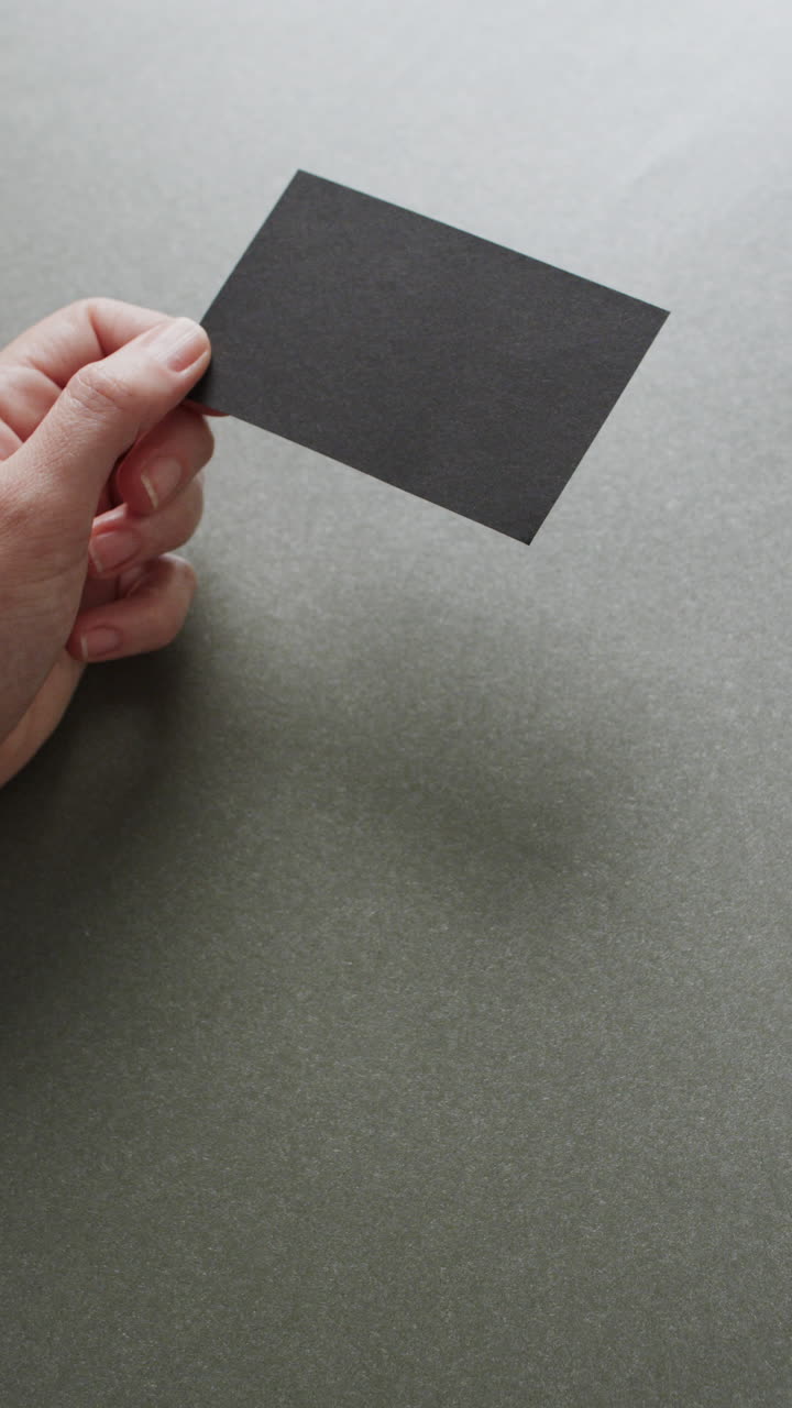 Vertical video of hand of caucasian woman holding black business card on grey background, copy space