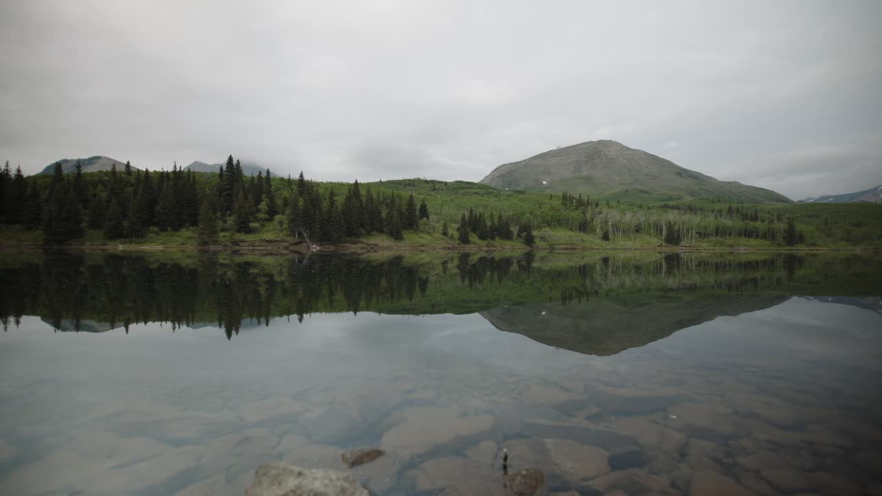 A tranquil mountain lake mirror reflects the surrounding forest and rolling hills under a soft overcast sky, showcasing natures stillness and the unspoiled wilderness