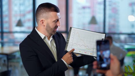 middle-aged employer speaks to the camera and shows the chart. hand holding phone taking video in blur at foreground.