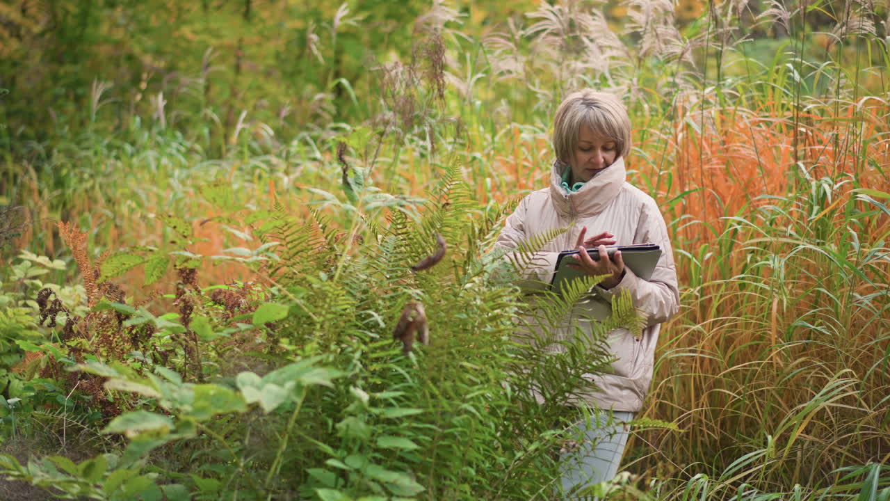 female researcher examining fern leaves in vibrant autumn vegetation while recording detailed observations on tablet device, surrounded by dense foliage in peaceful natural environment