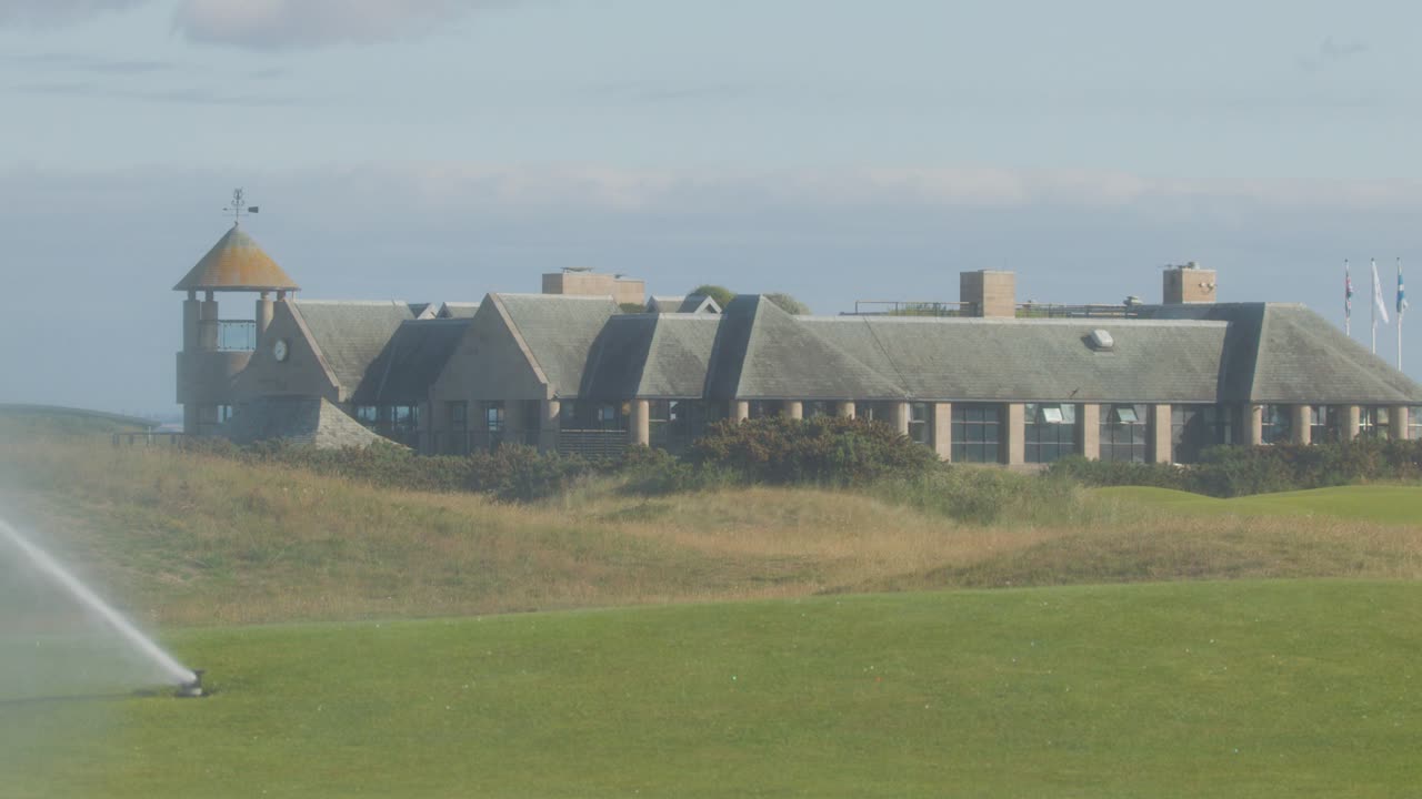 Wide shot of a golf course green being irrigated by a sprinkler, with a clubhouse in the background under natural daylight, steady camera