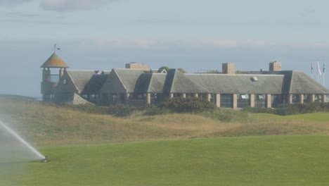 wide shot of a golf course green being irrigated by a sprinkler, with a clubhouse in the background under natural daylight, steady camera