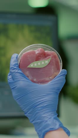 hand in blue glove holding a petri dish with 'vegan' labeled cultured meat