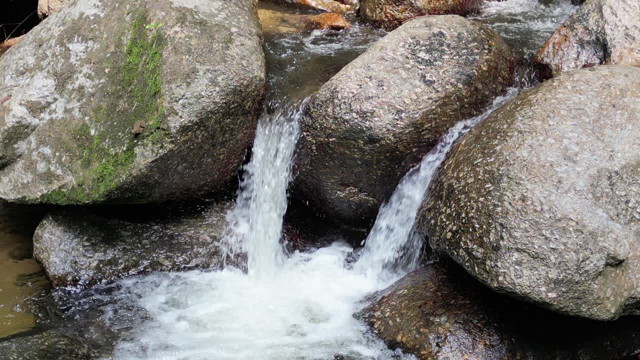 Cinematic close up view of small waterfall stream flowing between large rounded rocks with white water creating natural texture, gentle motion, and calm outdoor forest scenery
