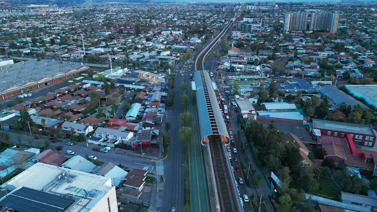 Rekaman Orbit Udara di Sekitar Kereta dengan Gerbong Penumpang Oranye yang Menyala di Rel Melintasi Area Perumahan Perkotaan dengan Lalu Lintas Padat di Jalan Raya Paralel saat Senja. Santiago, Chili