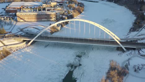 Winter-Aerial-birds-eye-view-dolly-roll-fly-over-the-North-Saskatchewan-River-covered-with-snow-ice-headed-towards-the-white-modern-walter-dale-bridge-as-sun-shines-on-the-bow-tied-arch-design-5-5