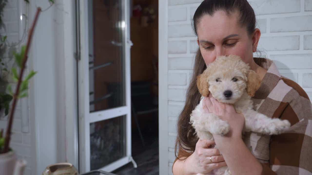 Woman in checkered shirt lovingly holding golden retriever puppy near face while sitting indoors, surrounded by houseplants and gardening items on table, enjoying peaceful bonding moment with pet