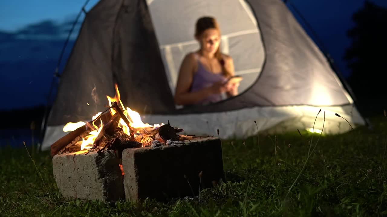 Bål med glødende flammer i forgrunden og kvinde, der sidder inde i telt og bruger smartphone, fanger hyggelig udendørs campingstemning og moderne livsstil i Savci, Slovenien