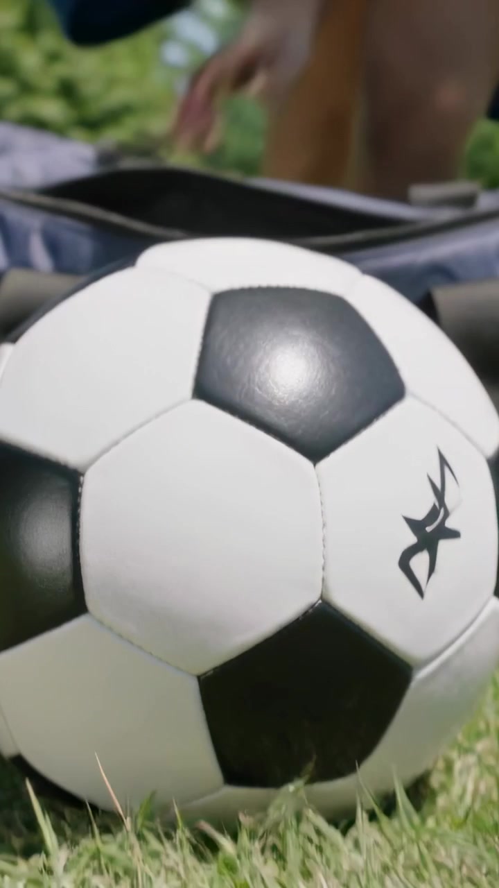 Dikey video: Çantanın fermuarını açan, tişörtlü iki çocuk futbol topunu çime çekiyor ve oynamaya hazırlanıyor.
