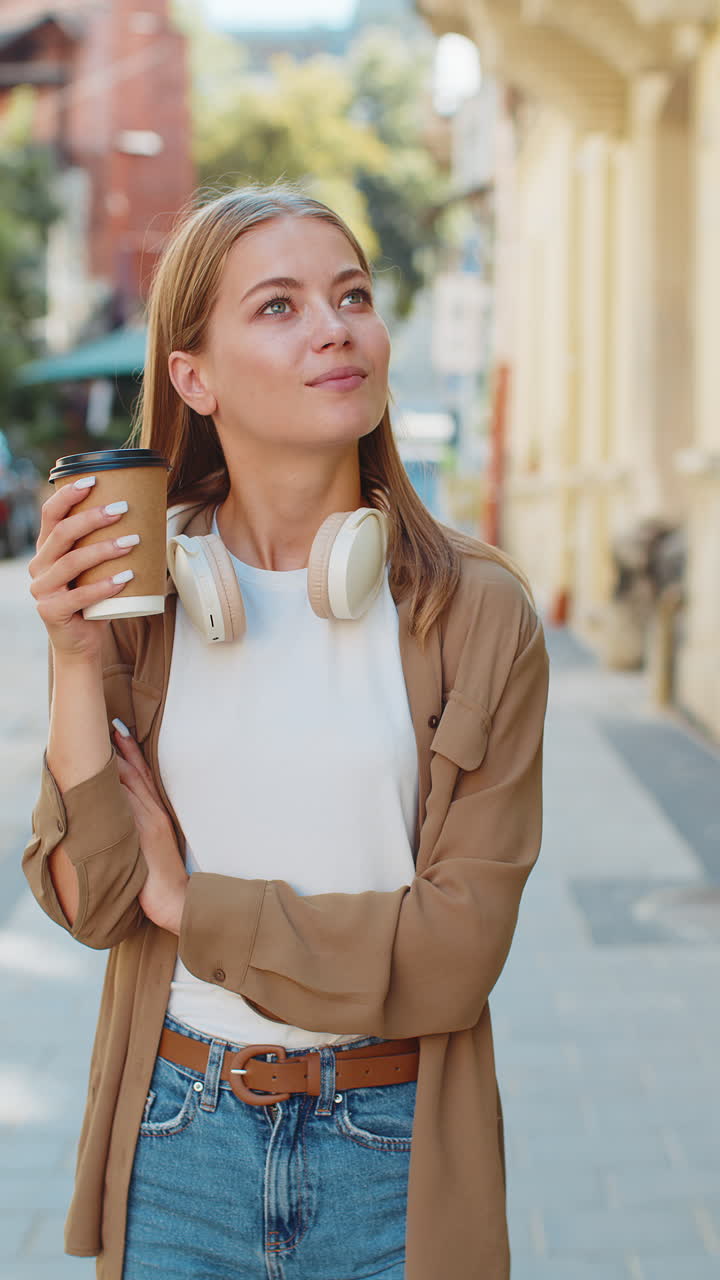 행복한 백인 젊은 여성이 아침 커피를 즐기고 뜨거운 음료를 마시며 웃으며 도시 거리를 고 있습니다.
