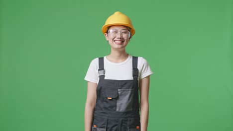 asian woman worker wearing goggles and safety helmet standing and smiling to camera in the green screen background studio