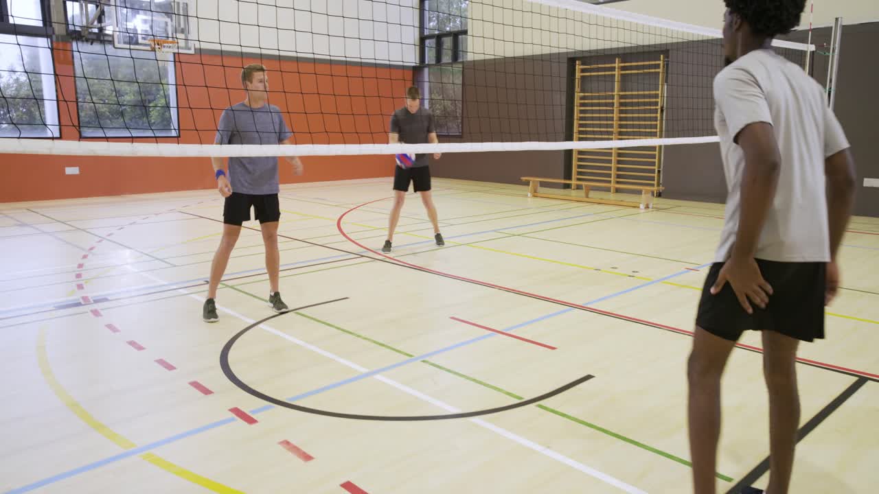 Pět mužů začíná volejbalovou výměnu ve školní tělocvičně, kudrnatý muž skáče, aby vrátil fialový volejbalový míč.