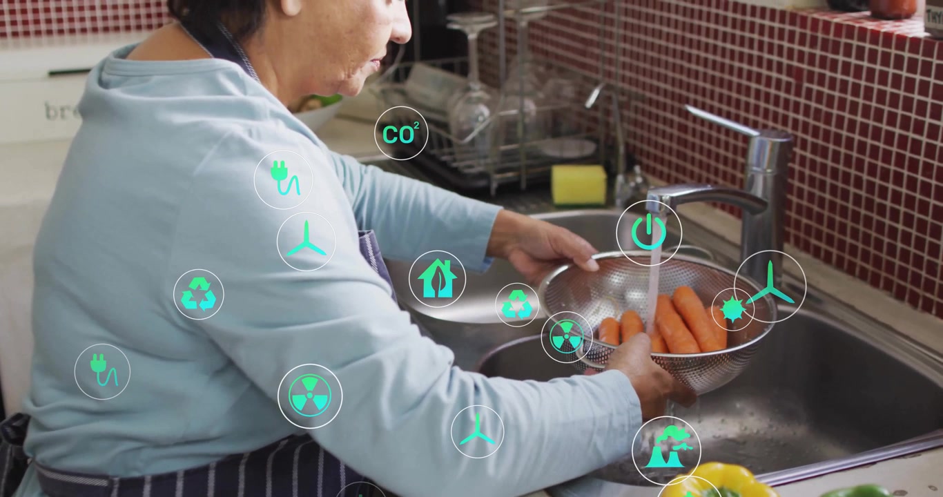Mujer mayor lavando zanahorias en el fregadero de la cocina, mostrando iconos de energía ecológica para educación sanitaria.