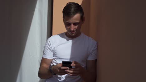 Happy-man-texting-on-a-smart-phone-outdoors-leaning-against-a-wall-with-the-sun-shinning-on-him