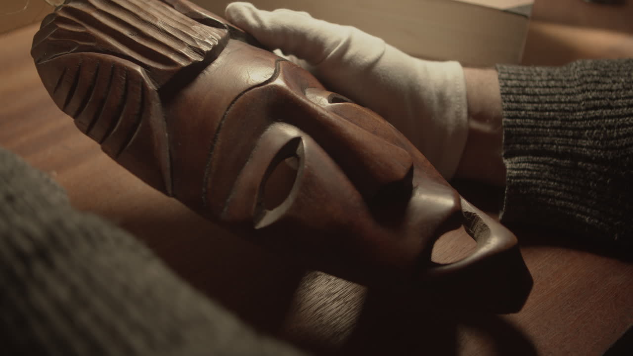 Curador com luvas a inspecionar uma máscara tradicional Baule ou Guro num ambiente de museu. Ideal para temas de património, conservação e exposição.