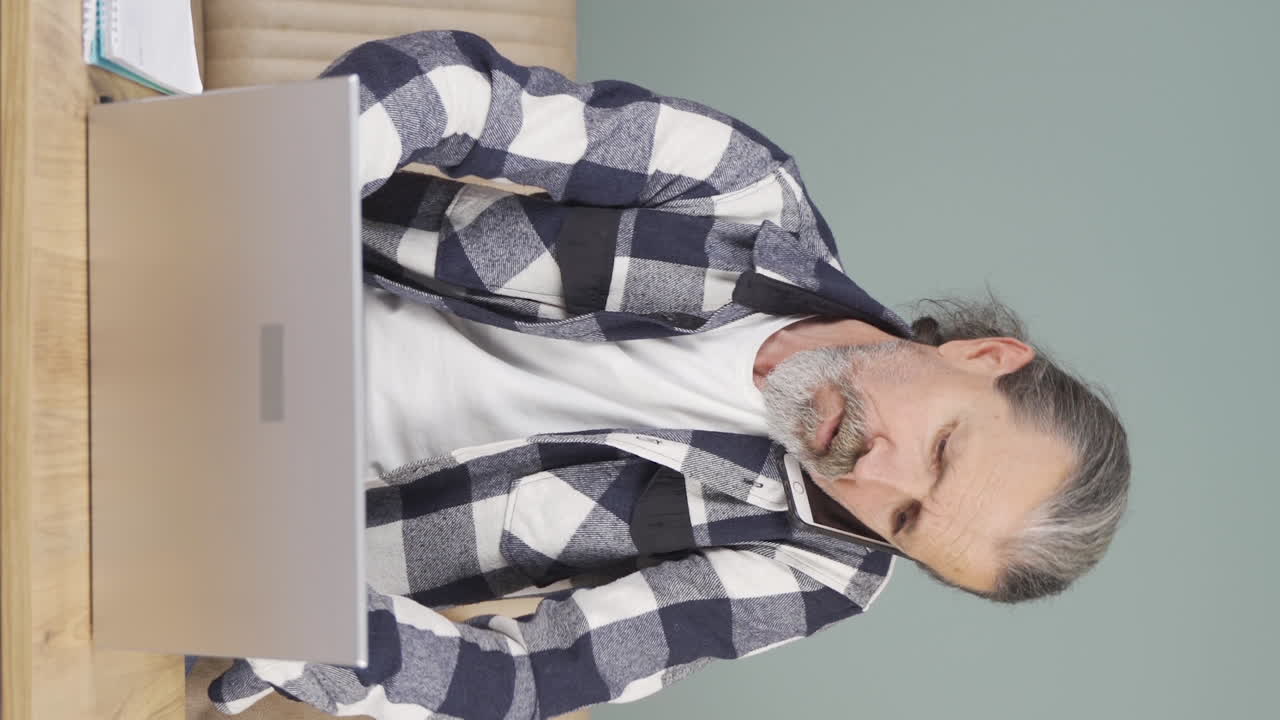 video vertical de un anciano usando una computadora portátil hablando nerviosamente por teléfono.