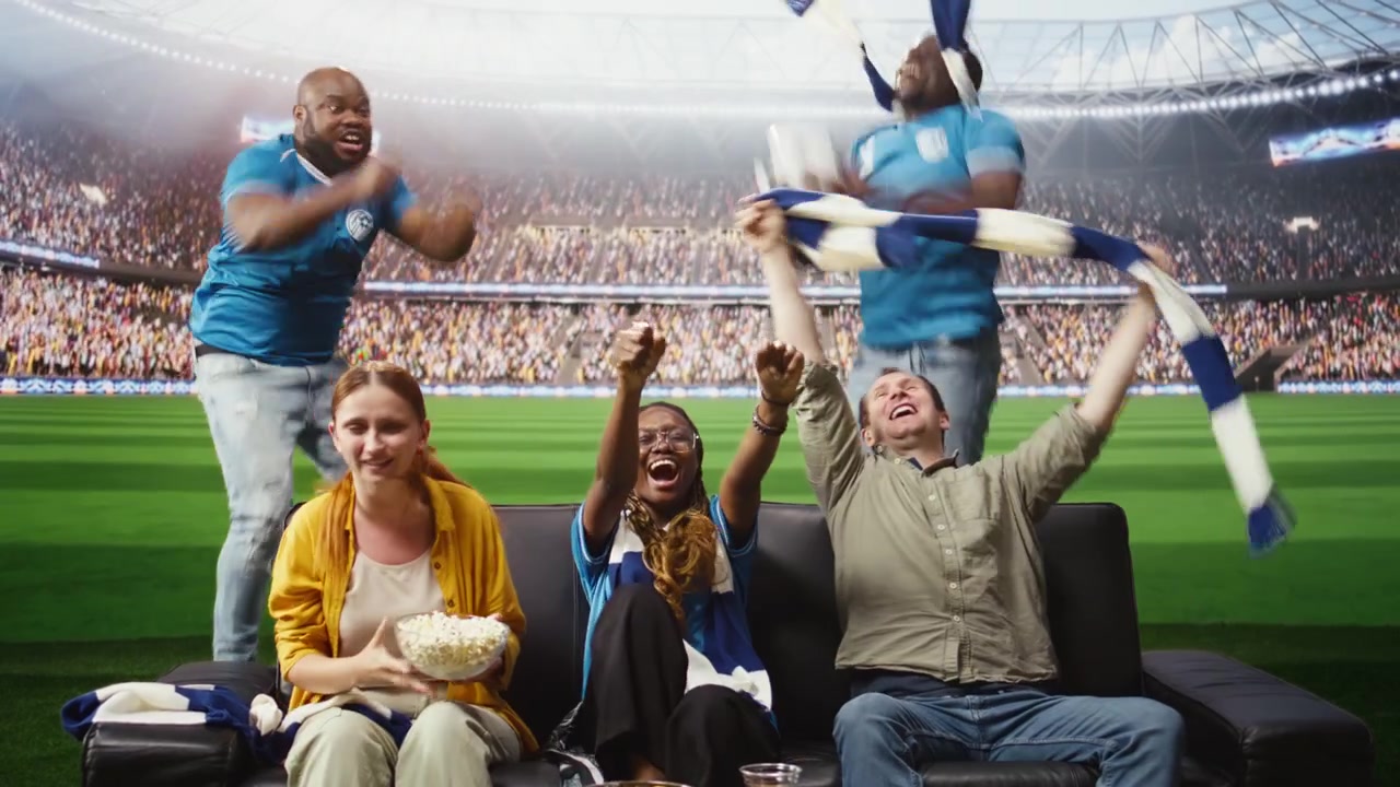 Alegres aficionados al fútbol en el sofá animando después de un gol, sintiéndose como si estuvieran dentro del estadio.
