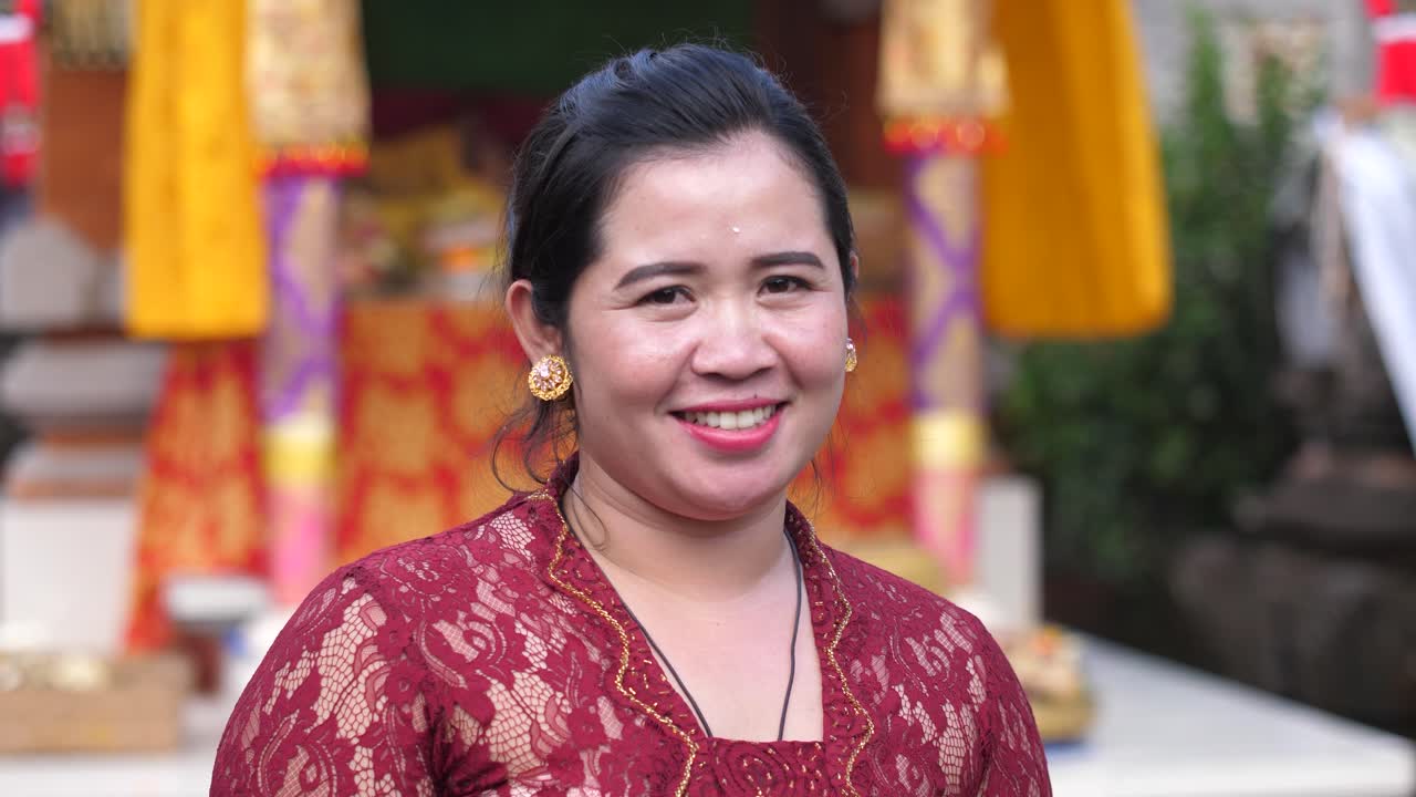 Mulher feliz com vestido de renda vermelho escuro de pé em frente a cortinas e faixas coloridas em um templo hindu em Bali