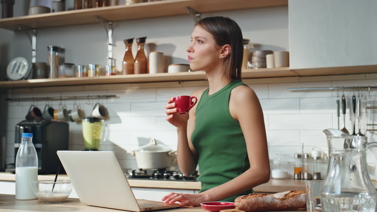 Woman working on laptop in kitchen while drinking coffee
