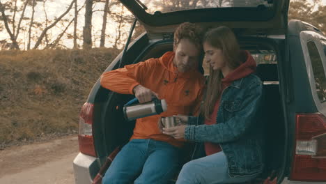 -Young-woman-and-man-sitting-in-the-open-trunk-of-the-car-having-a-hot-drink-with-a-thermos