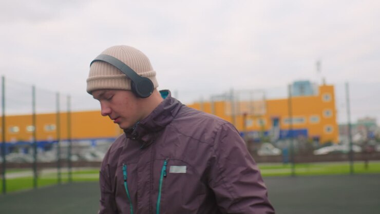 White man practicing basketball with headphones on urban outdoor court wearing beanie and jacket warming up taking shots listening to playlist focused expression cloudy sky yellow building fence