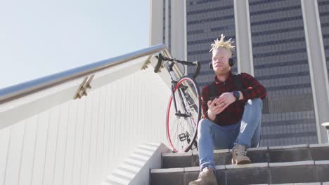 Thoughtful-albino-african-american-man-with-dreadlocks-wearing-headphones-using-smartphone