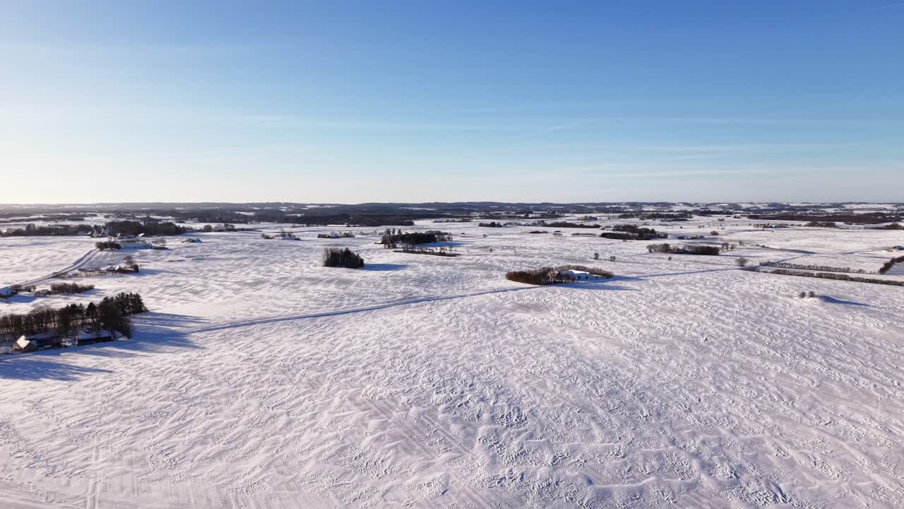 Cinematic drone pan over a serene, snow-covered countryside. This high-angle view captures frozen agricultural fields, distant farmhouses, and a clear blue horizon during a bright winter day