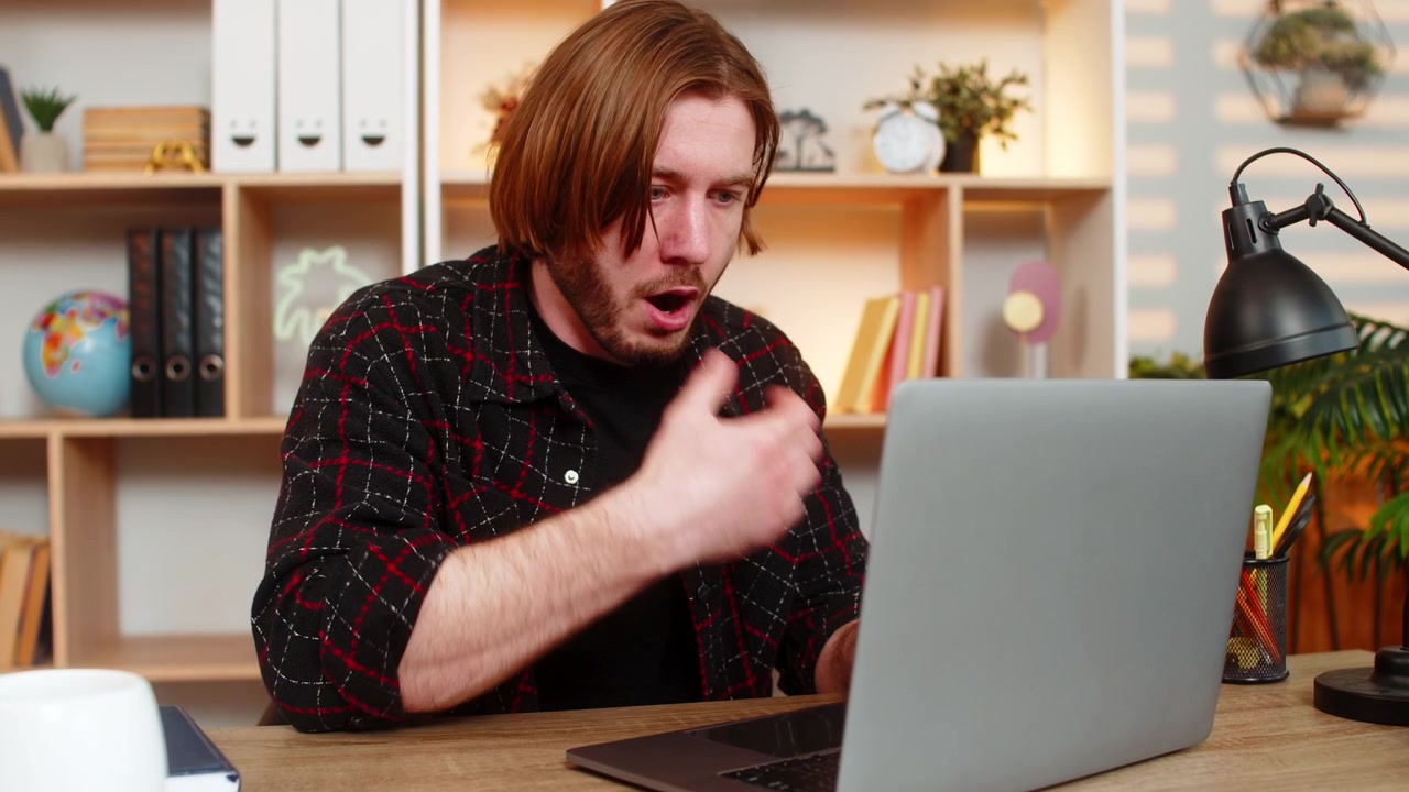 Young man on home table reacting to important news on laptop screen with surprise during remote work