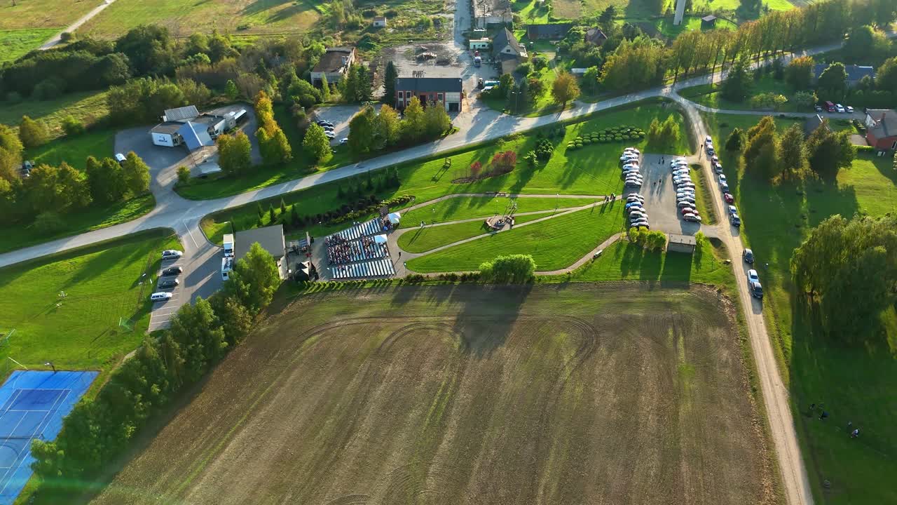 Aerial view of an outdoor stage and seating area set within a green suburban park, surrounded by trees, roads, and open fields in warm evening light