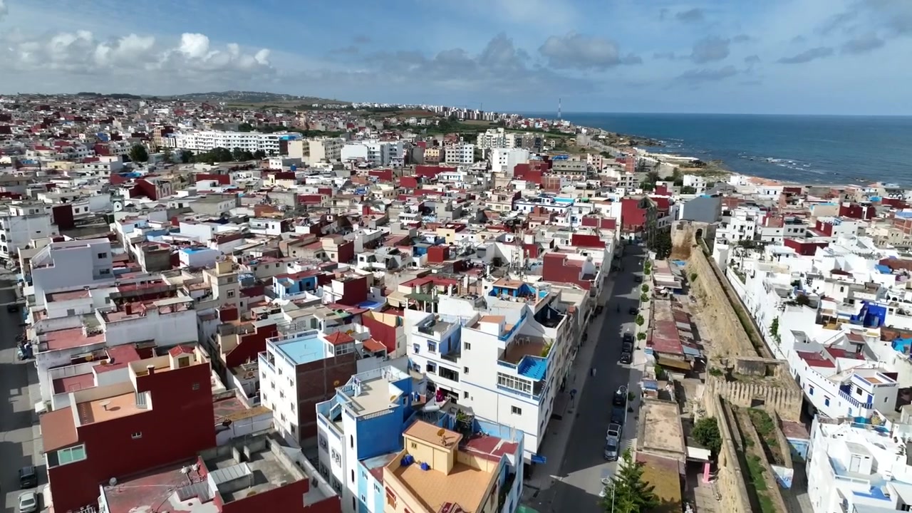 Bredt luftfoto af Asilah, Marokko, der viser tætte hvide og farverige kvarterer, en lang bygade, historiske befæstede mure og Atlanterhavskysten i baggrunden.