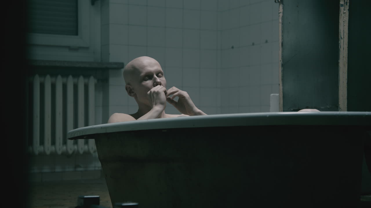 Pale bald skinny man takes a bath in the creepy old bathroom at night