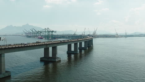 Panorámica-Aérea-Que-Revela-La-Vista-De-Los-Vehículos-Que-Conducen-En-El-Tráfico-Sobre-La-Bahía-De-Guanabara-En-El-Puente-De-Rio-niteroi-Que-Cruza-El-Mar-Con-Vista-Al-Puerto-Marítimo