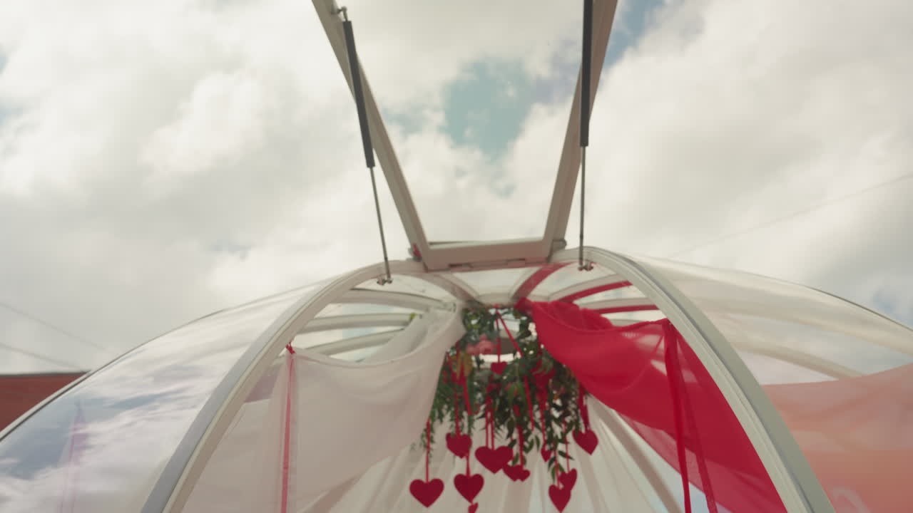 baldacchino sul tetto con drappeggio rosso e bianco che incornicia cuori e vegetazione sospesi, tulle trasparente che si gonfia nella brezza, centrotavola con lampadario floreale, sfondo di cielo nuvoloso, ambiente intimo e romantico perfetto