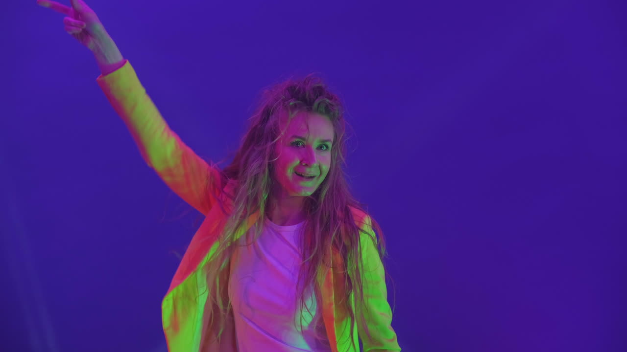 The woman dances merrily and looks at the camera in the light of strobe lights and spotlights. Neon colors and crazy girl dancing to music and singing