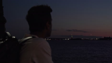 steady medium shot at night of a man gazing over the hudson river from manhattan pier, illuminated by the soft city lights. the focus shifts towards the statue of liberty in the distance.